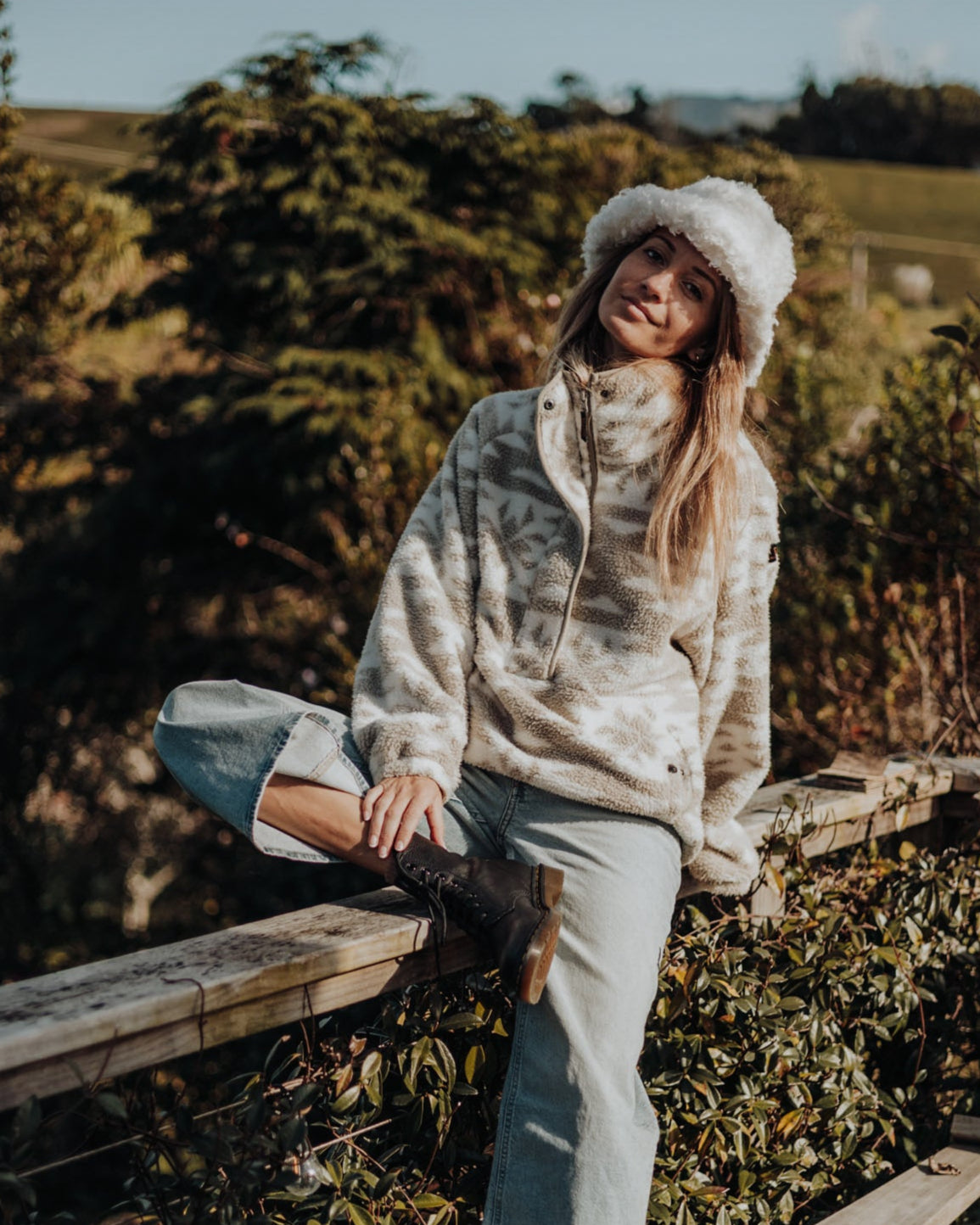 Woman sitting on a wooden bench in a natural setting with trees and open sky.