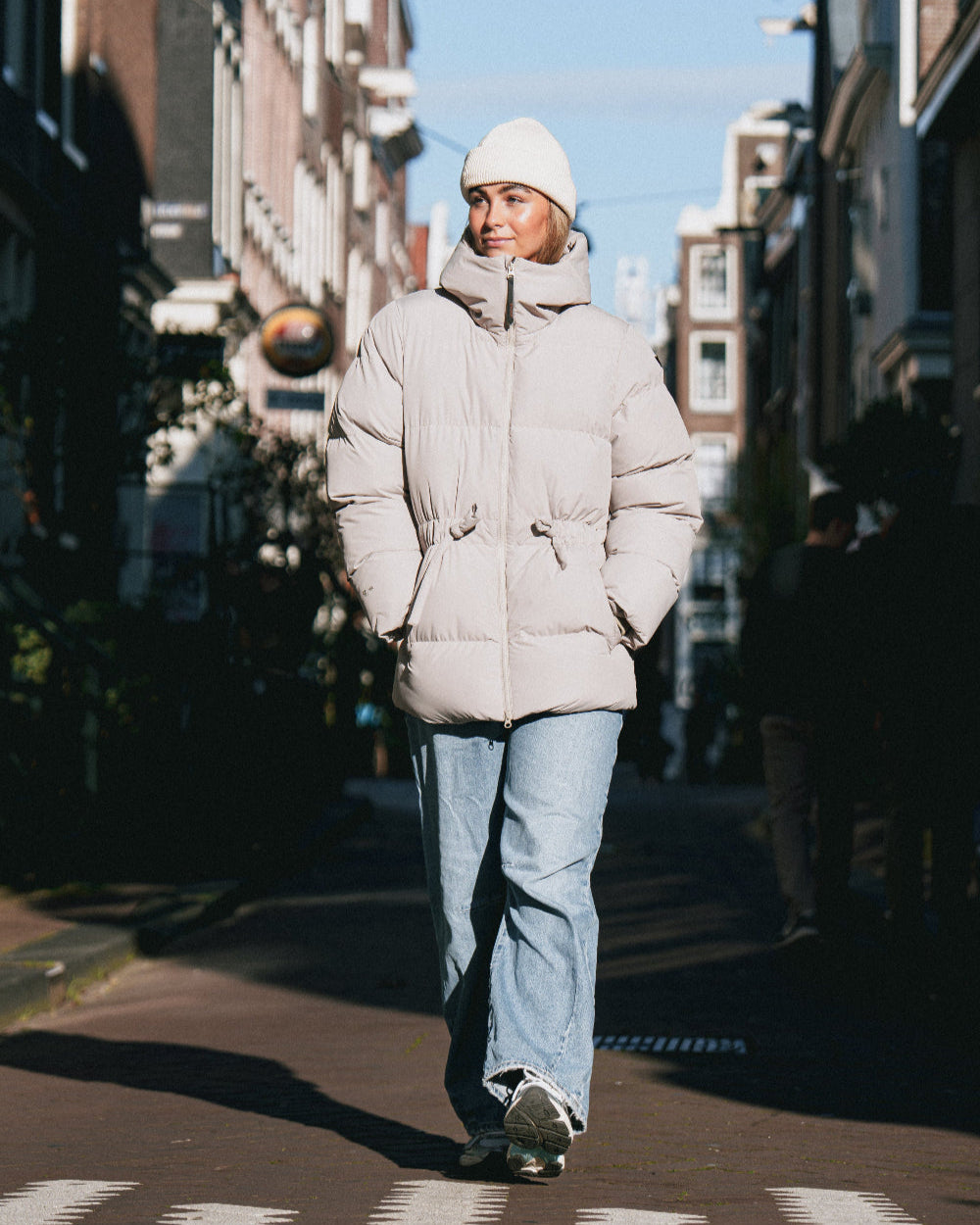 Person wearing a light gray puffer jacket and blue jeans walking on a city street.