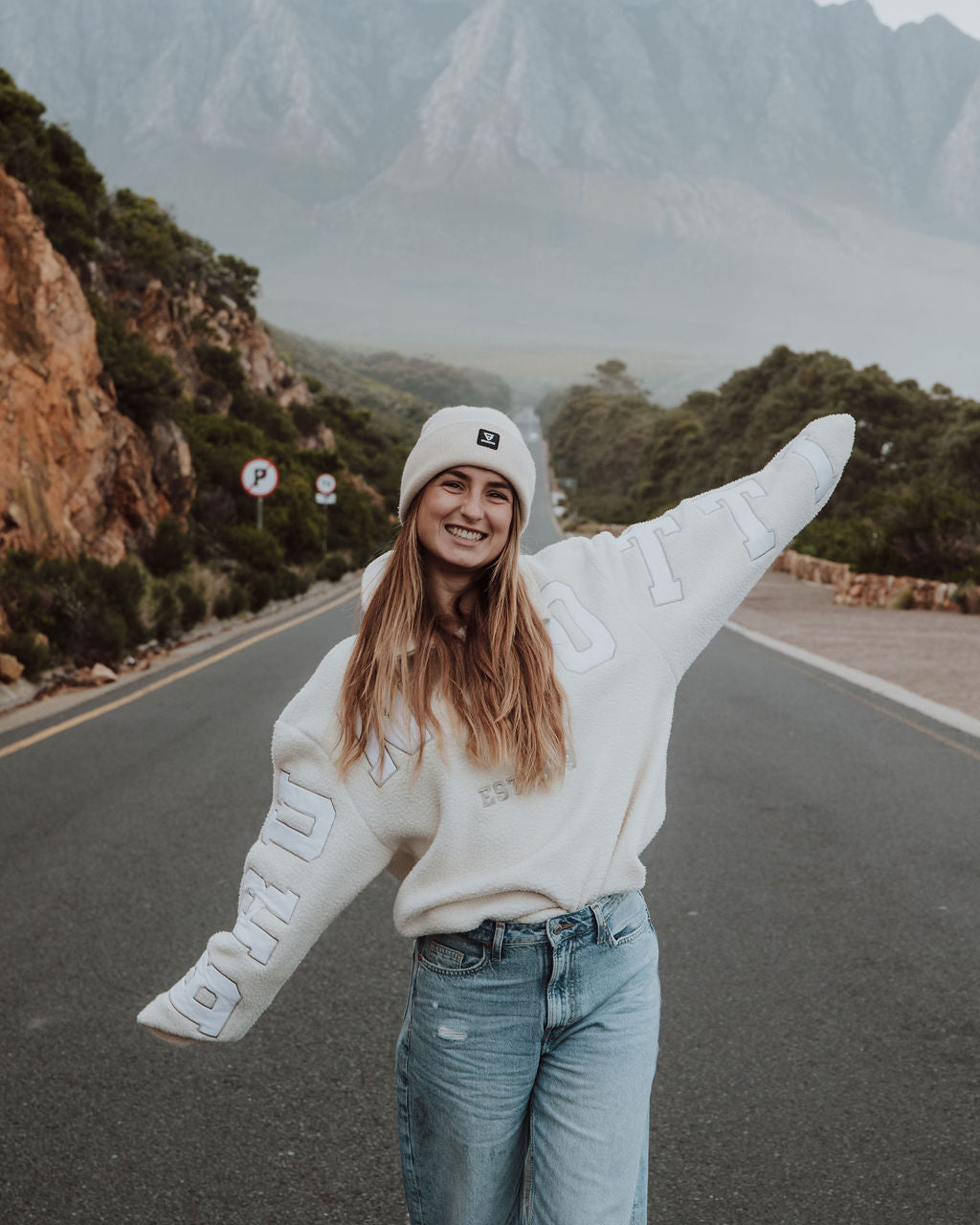 Person in a white sweater and beanie standing on a road with mountains in the background