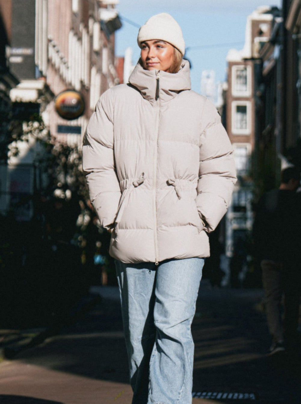 Person wearing a beige puffer jacket and light blue jeans walking on a street.