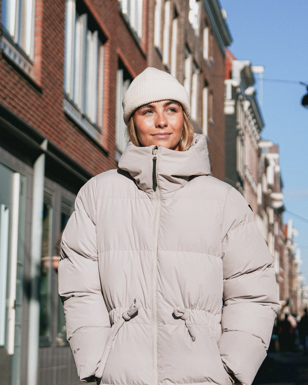 Person wearing a light gray puffer jacket and white beanie standing on a city street.