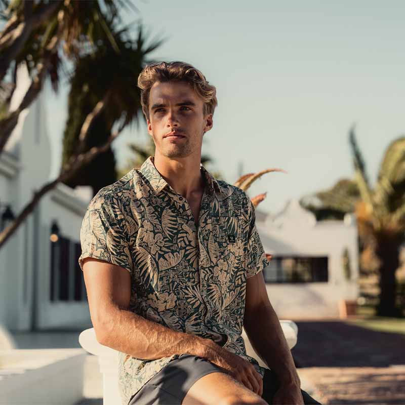 Jason van der Spuy wearing the new brunotti Summer collection with a beige and green shirt with short sleeves and a grey shorts. He is sitting in the sun in Summer time.