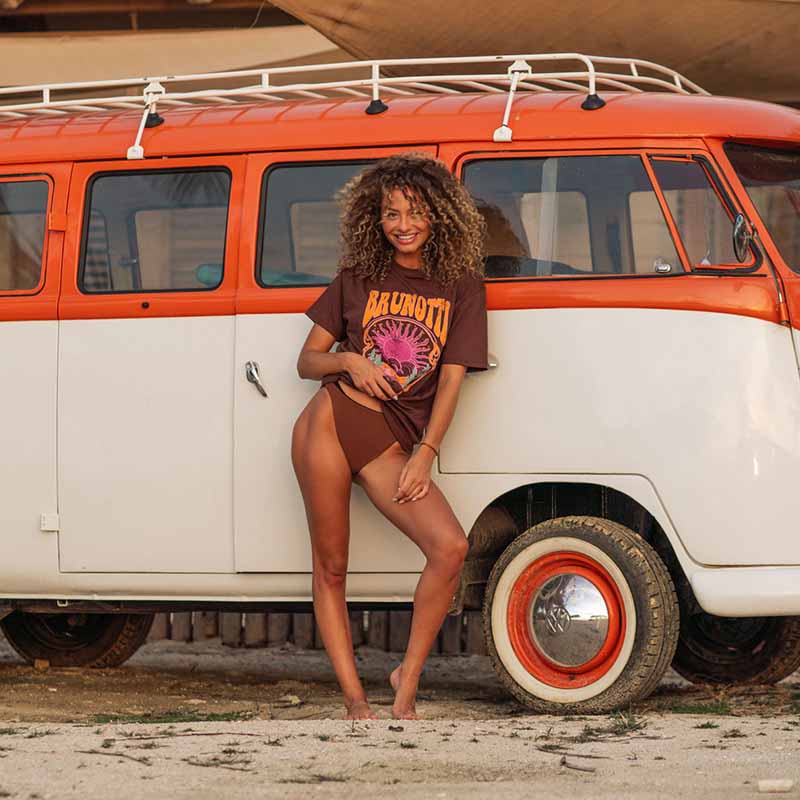 Smiling girl wearing the new Brunotti Summer Collection with a brown Brunotti T-Shirt and a brown Brunotti bikini bottom. Standing on the beach infront of an orange Camper van.